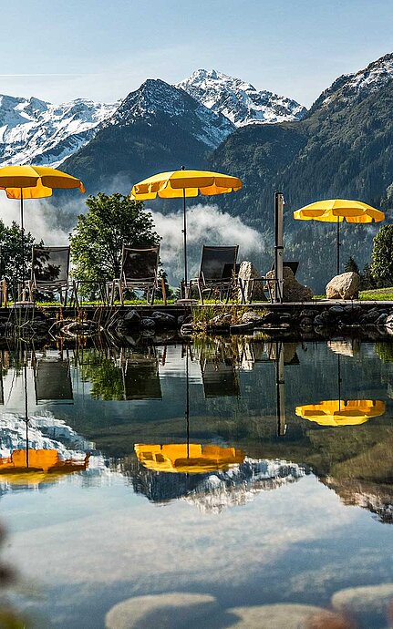 Hotel mit Badeteich im Salzburger Land mit gelben Sonnenschirmen und majestätischen Alpen im Hintergrund
