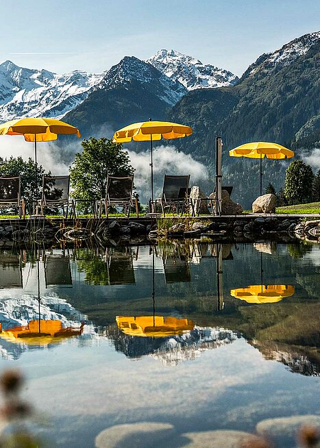 Hotel mit Badeteich im Salzburger Land mit gelben Sonnenschirmen und majestätischen Alpen im Hintergrund