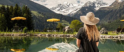 Frau in Sommerkleid und Strohhut vor Badeteich genießt die Aussicht auf die Berge im Naturhote