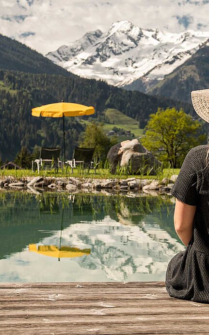Frau in Sommerkleid und Strohhut vor Badeteich genießt die Aussicht auf die Berge im Naturhote